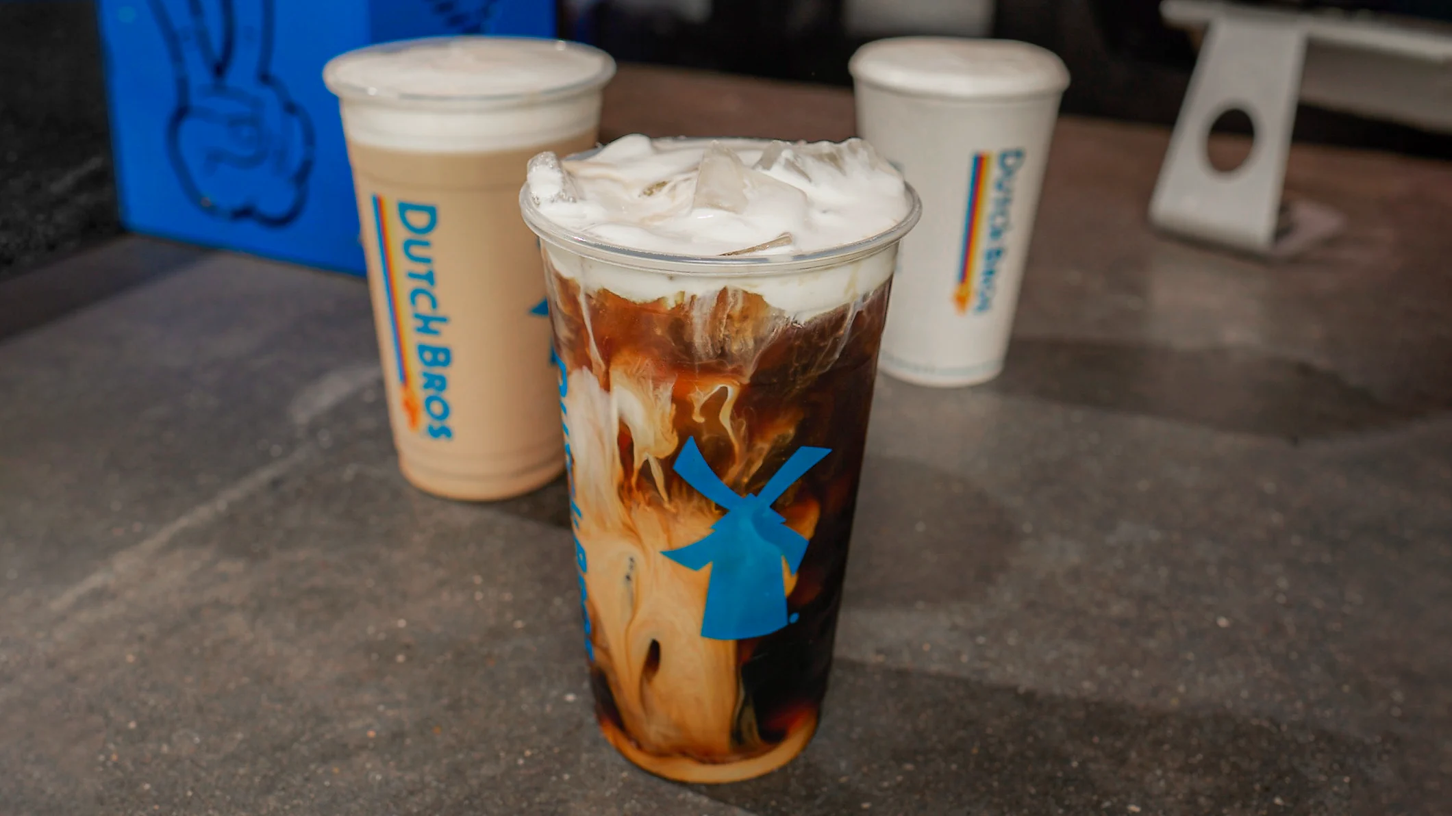 Three Dutch Bros coffee drinks on a counter - iced coffee with cream swirls in front and two light-colored drinks behind.