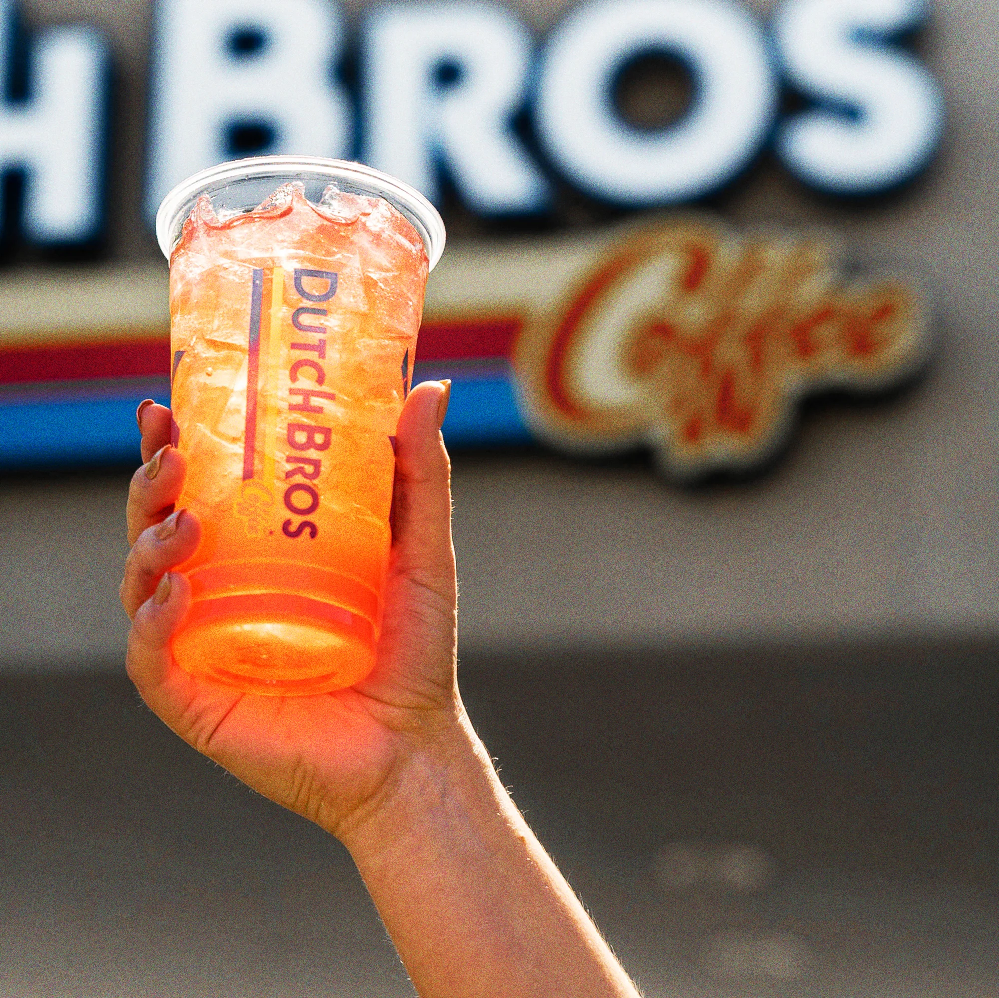 Hand holding an orange Dutch Bros iced drink with a blurred fast food restaurant storefront in the background.
