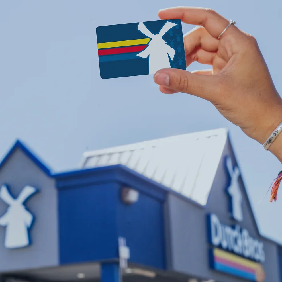 A hand holds a Dutch Bros gift card featuring a windmill logo in front of a blue Dutch Bros coffee shop.