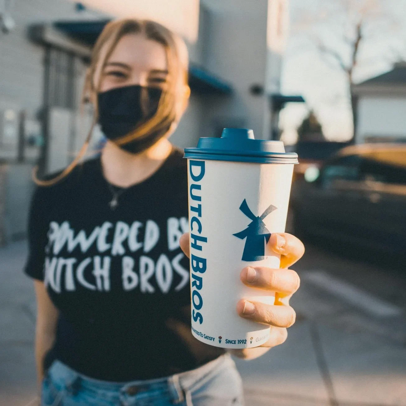 Person wearing black mask and shirt holding a Dutch Bros coffee cup with blue lid and windmill logo.