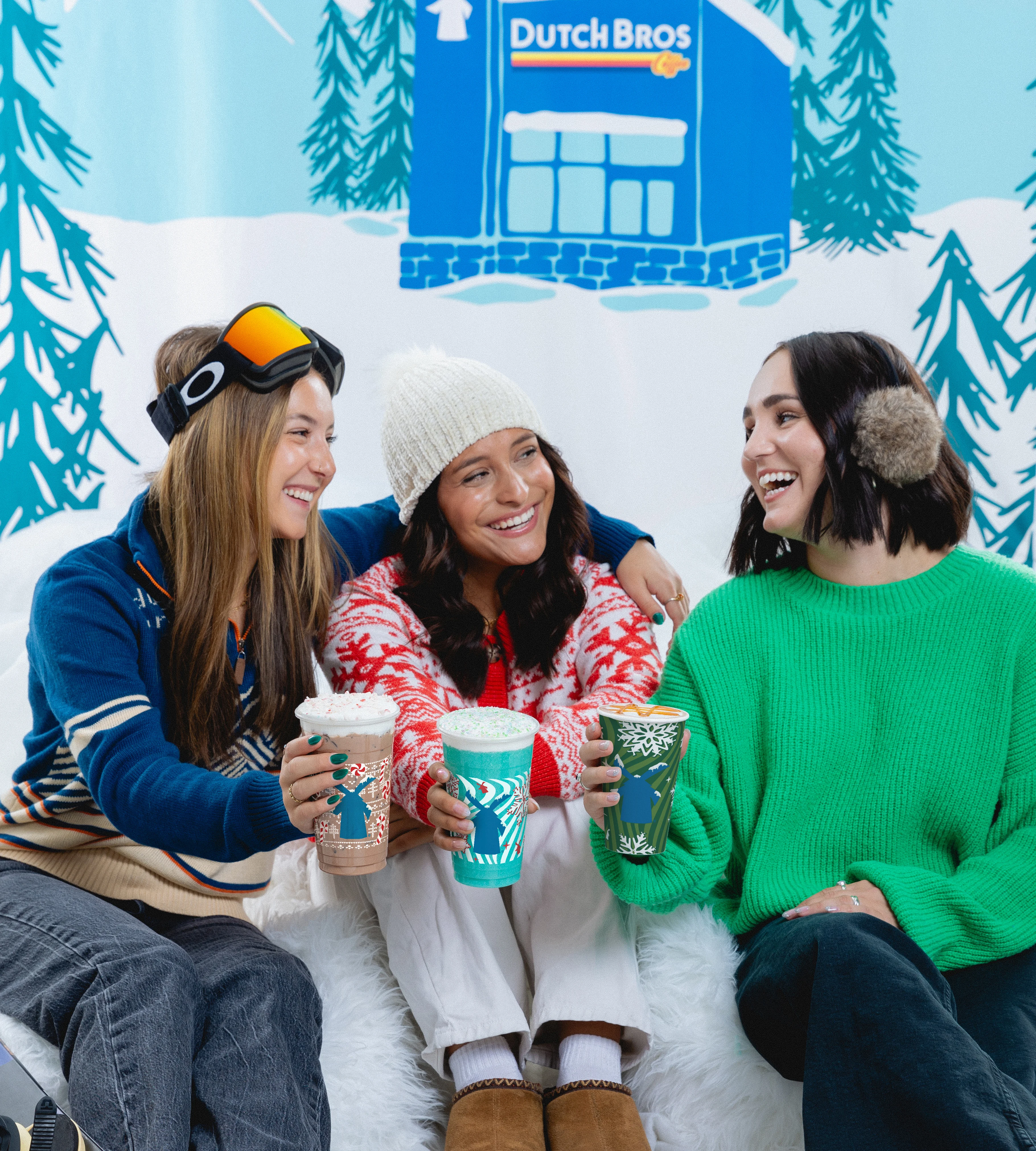 Three friends in winter attire enjoying Dutch Bros drinks, sitting against a snowy backdrop with pine trees.