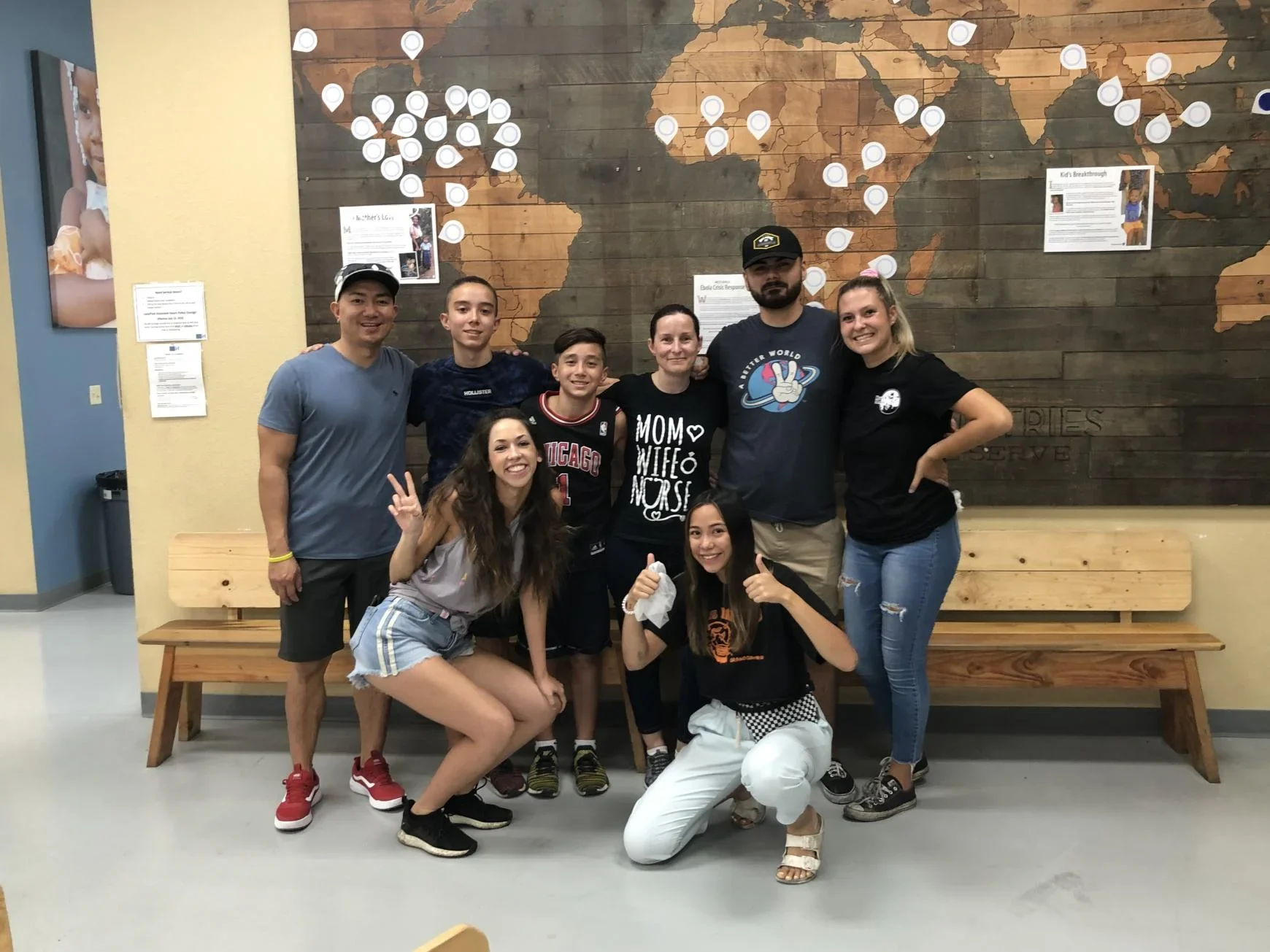 Group of people posing in front of a wooden world map with location markers, standing by wooden benches.