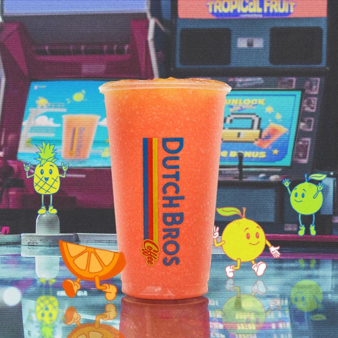 Orange smoothie in Dutch Bros cup with animated fruit characters and tropical fruit machine display in background.