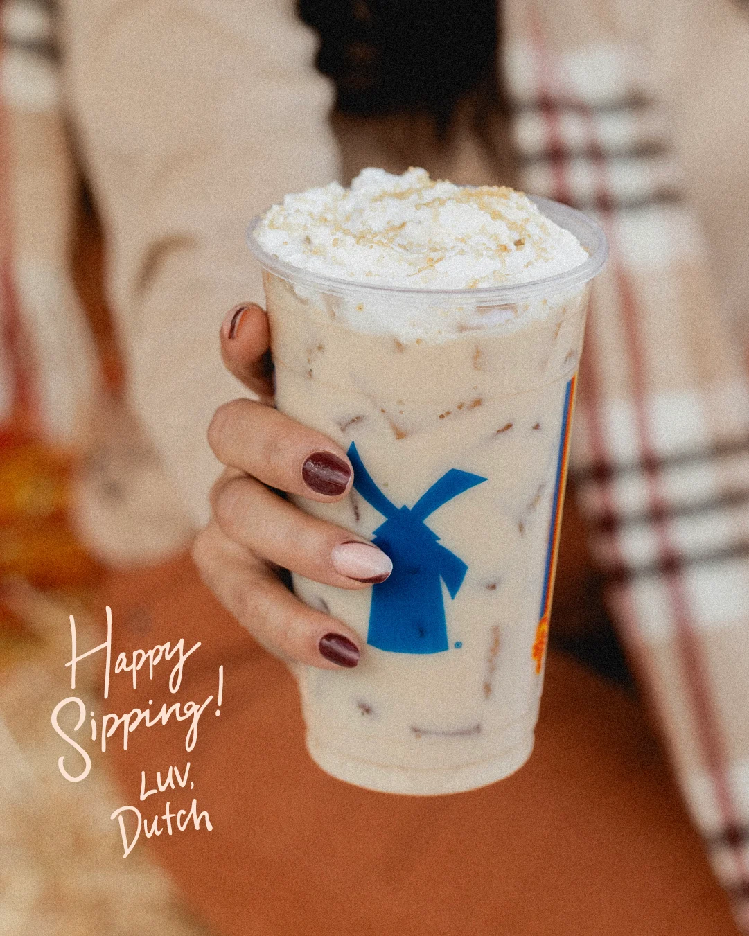 Hand with burgundy nails holding Dutch Bros iced coffee with whipped cream, text reads "Happy Sipping! Luv, Dutch"