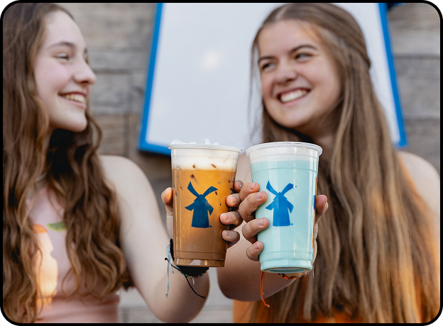 Two smiling friends toasting with colorful Dutch Bros drinks, one iced coffee and one blue beverage with windmill logos.