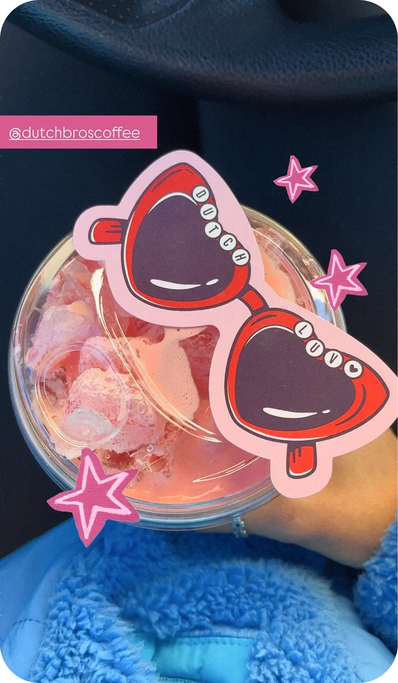A pink Dutch Bros iced drink held in hand, overlaid with heart-shaped sunglasses stickers reading "Dutch Luv" and pink star graphics.