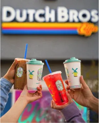 Hands holding three Dutch Bros coffee drinks with straws in front of the colorful Dutch Bros logo sign.