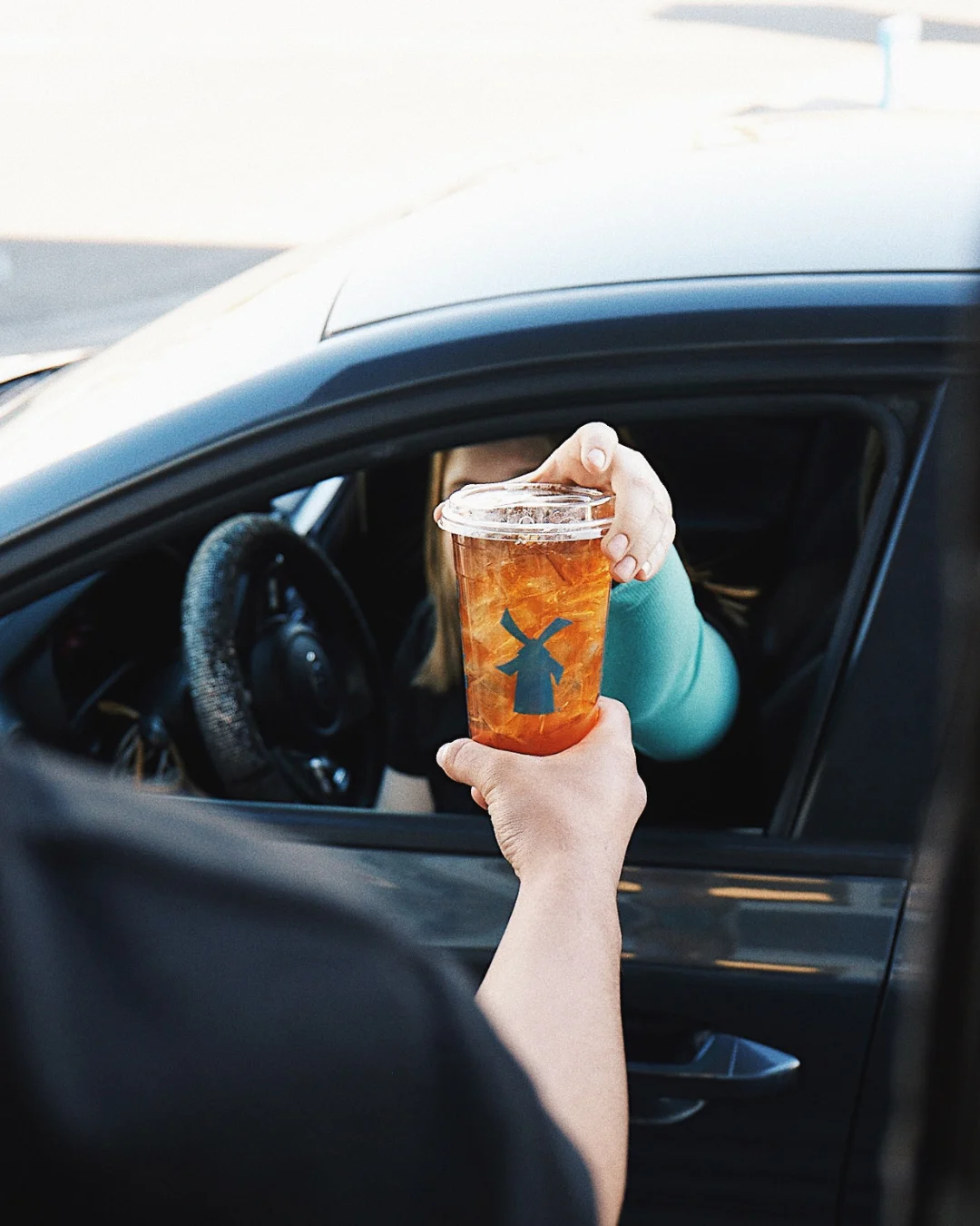 Hand passing an iced tea with blue windmill logo to someone in a car through the driver's window.