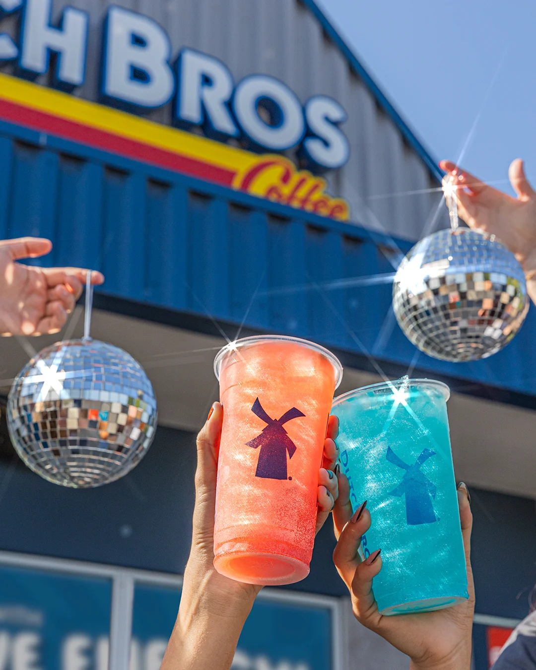 Hands holding colorful orange and blue drinks with windmill logos outside Dutch Bros Coffee shop with disco balls overhead.