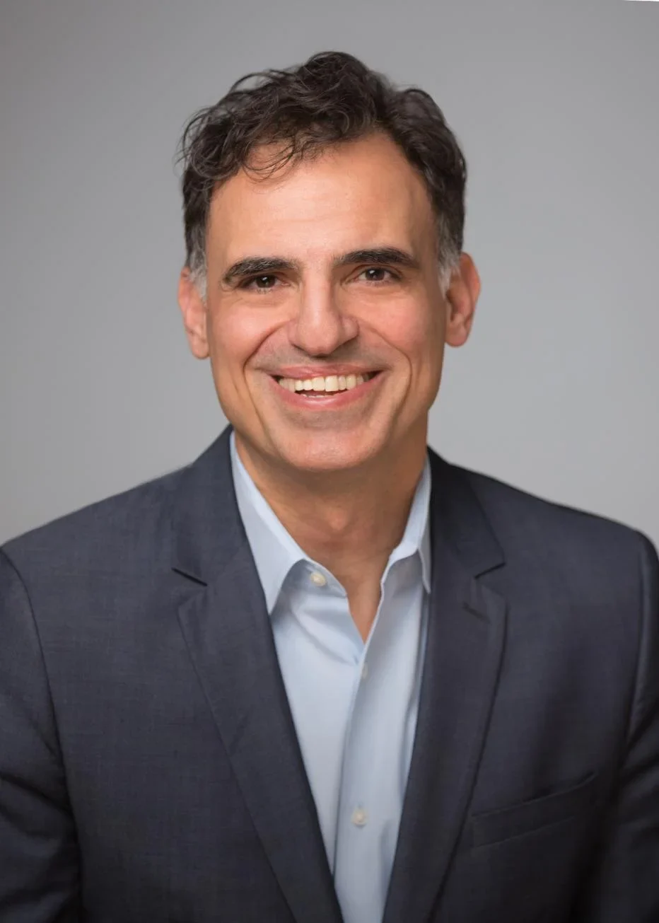 Professional portrait of a person with dark wavy hair wearing a navy suit and light blue shirt, smiling against a gray background.