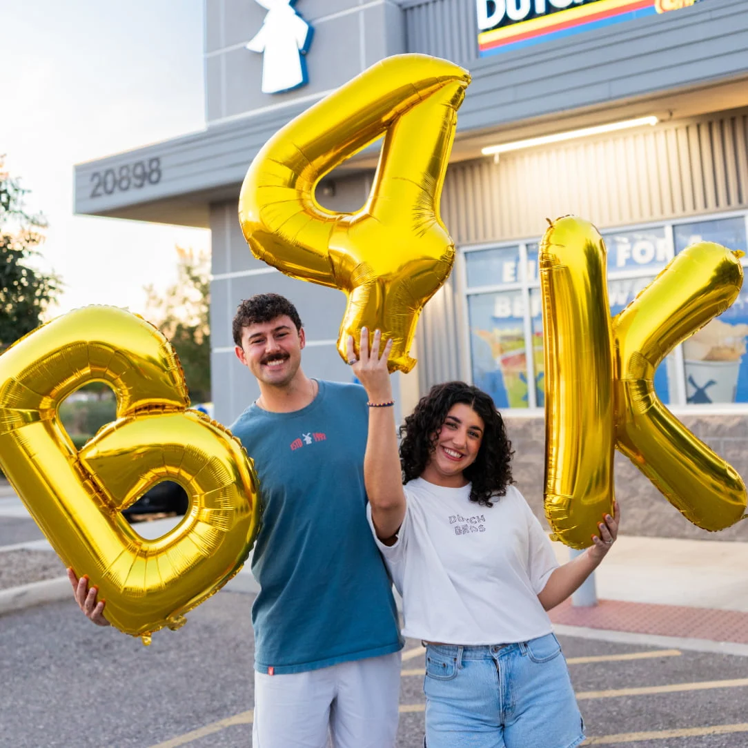 Two smiling people holding large gold balloon letters spelling "B4K" outside a storefront.