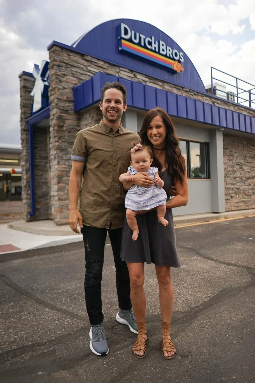 Family standing outside a Dutch Bros Coffee shop with blue awning and stone facade under cloudy sky.