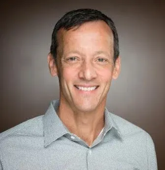 Professional headshot of a smiling person with short dark hair wearing a light patterned shirt against a brown background.