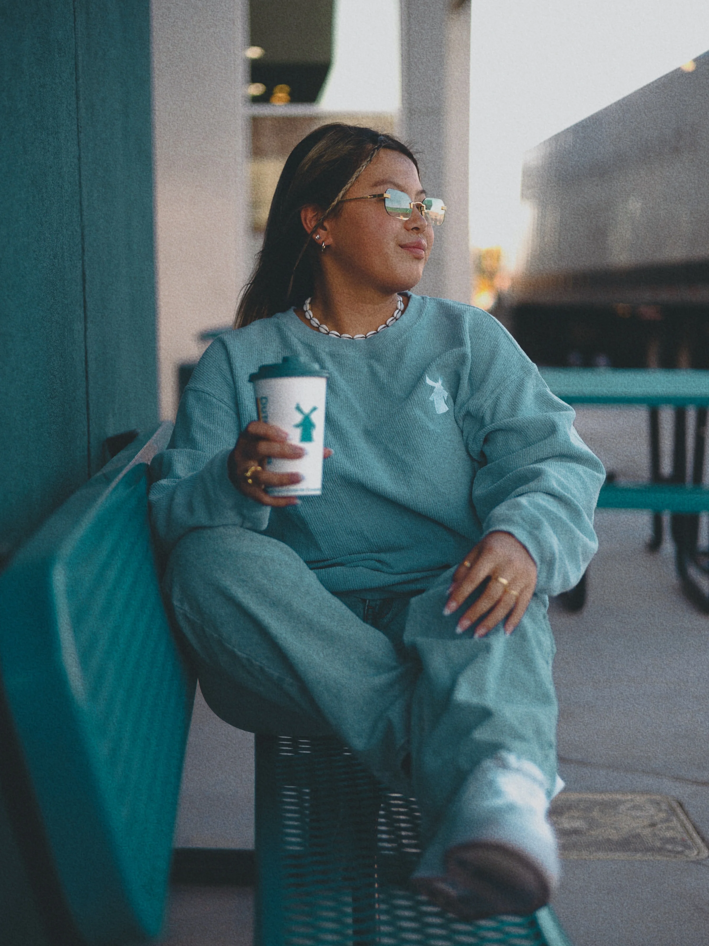 Person in blue sweatshirt with glasses holding coffee cup, relaxing on teal bench in modern cafe setting.