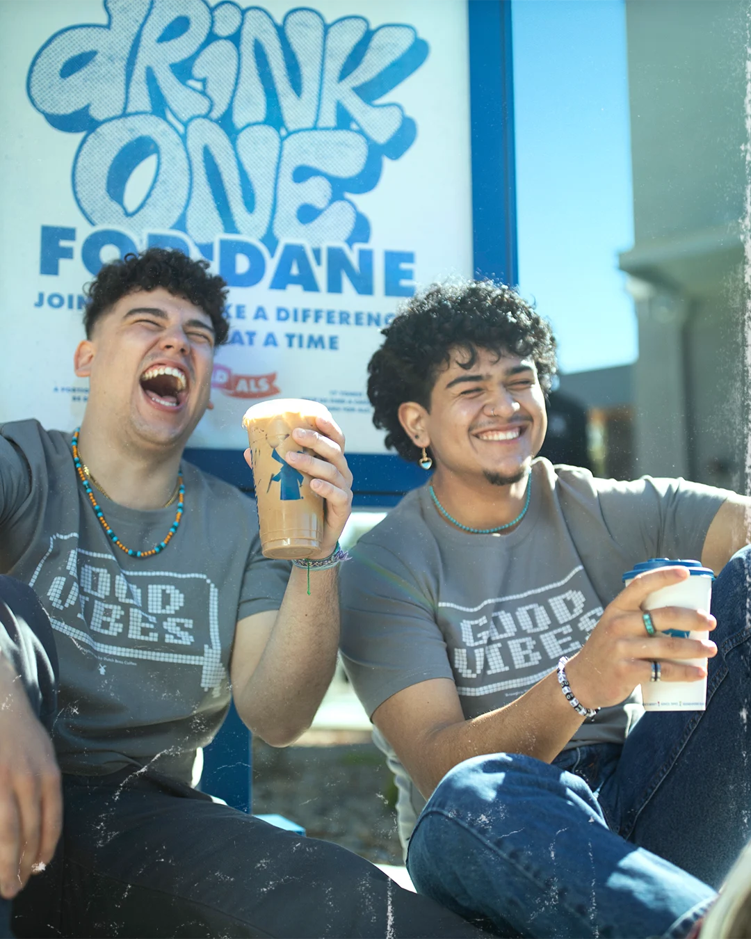 Two young people laughing while holding drinks, wearing matching "Good Vibes" t-shirts in front of a "Drink One" sign.
