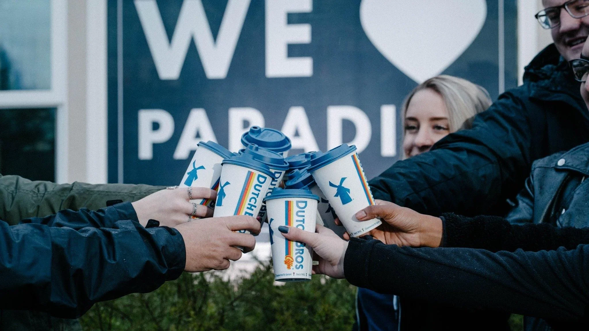Group toasting with Dutch Bros coffee cups in front of a blue "WE ♥ PARADI" sign.