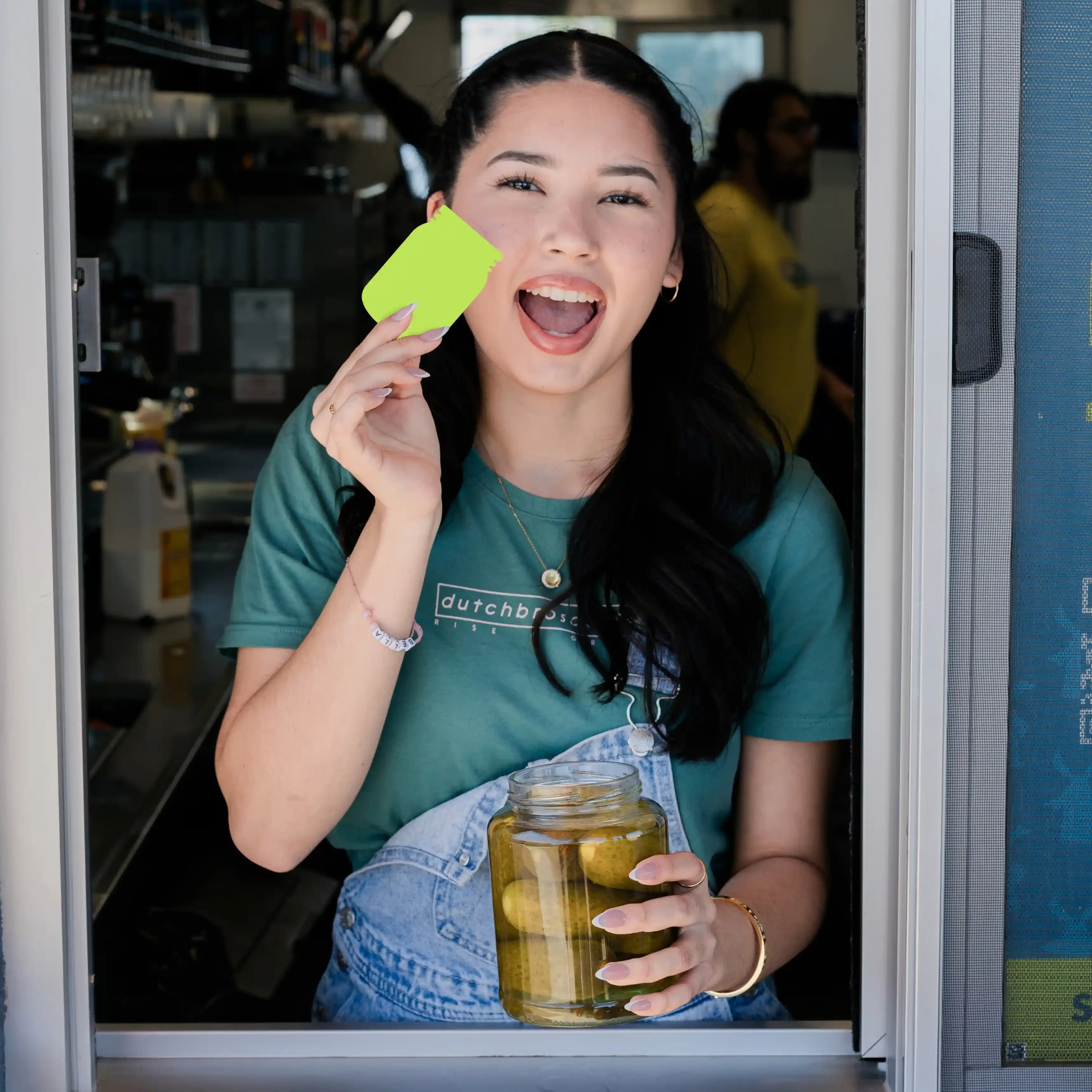 Broista in teal shirt holding bright green sticker and jar of pickles, smiling excitedly at shop window.