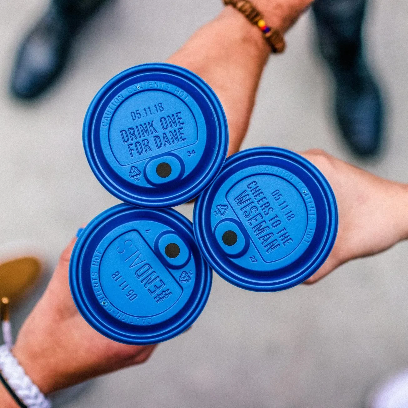 Hand holding three blue coffee cup lids with personalized messages including "Drink One For Dane" dated 05.11.18.