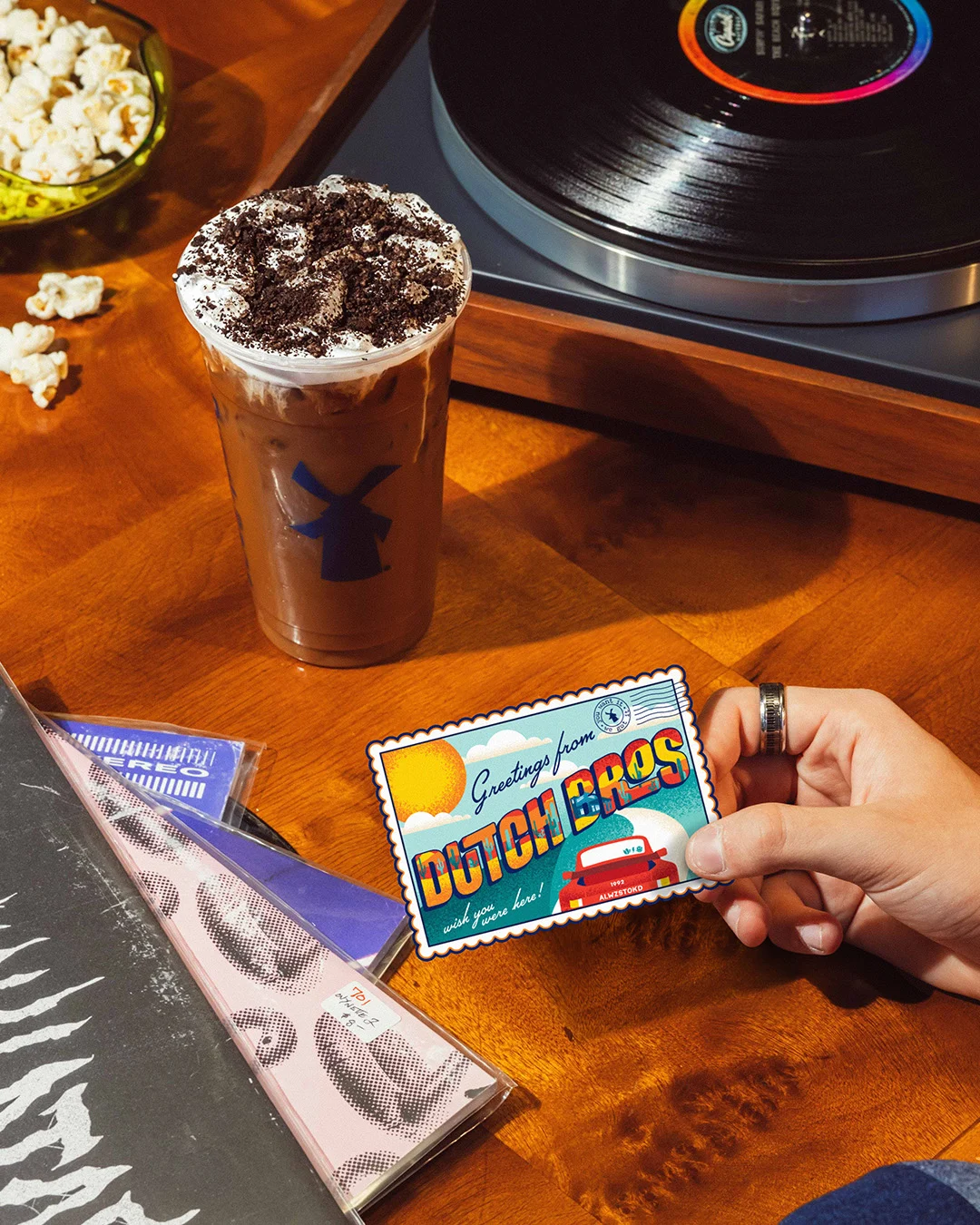 Iced coffee with whipped cream beside vinyl record player, popcorn bowl, and hand holding "Dutch Bros" postcard.