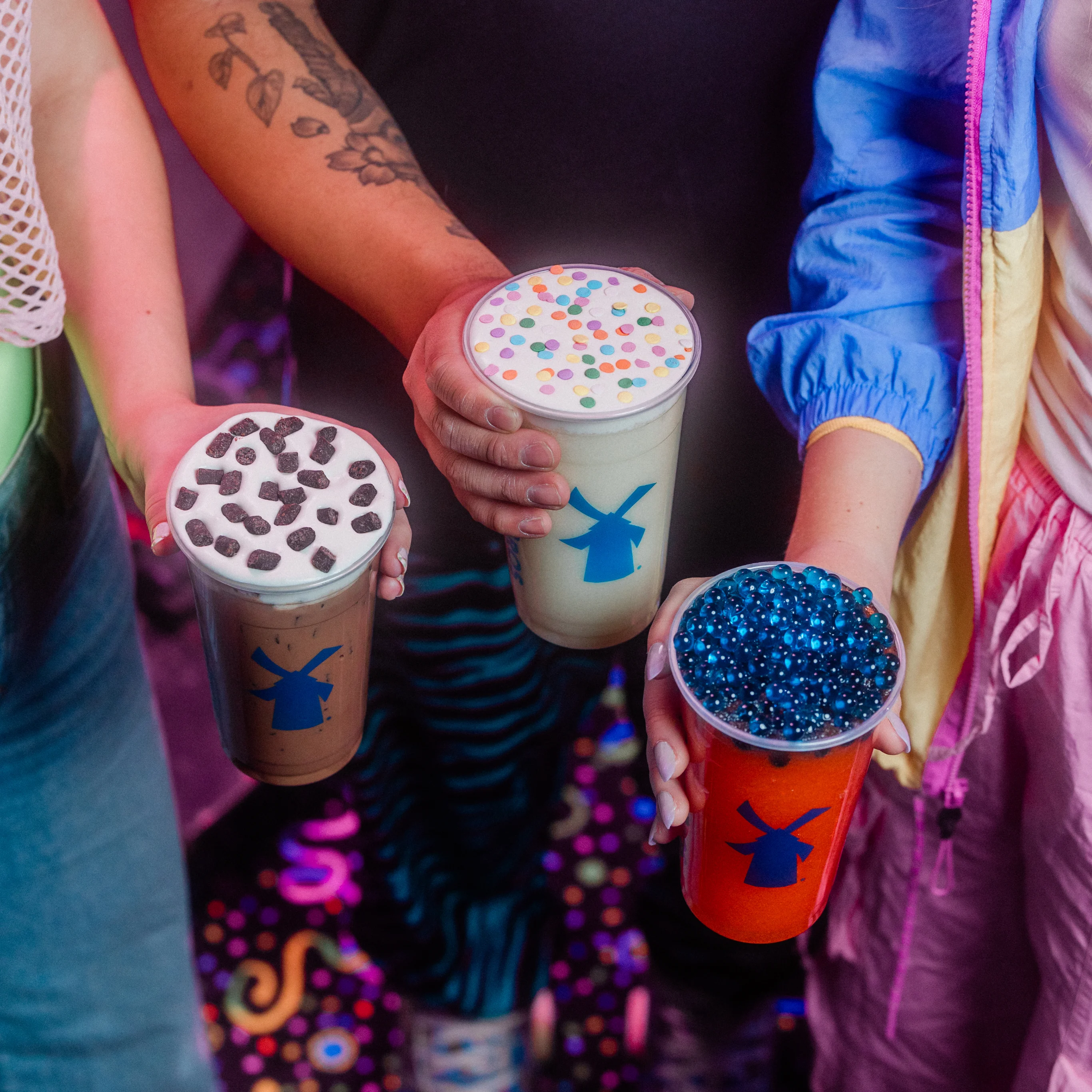 Hands holding three colorful bubble tea drinks with different toppings - chocolate pieces, rainbow sprinkles, and blue pearls.