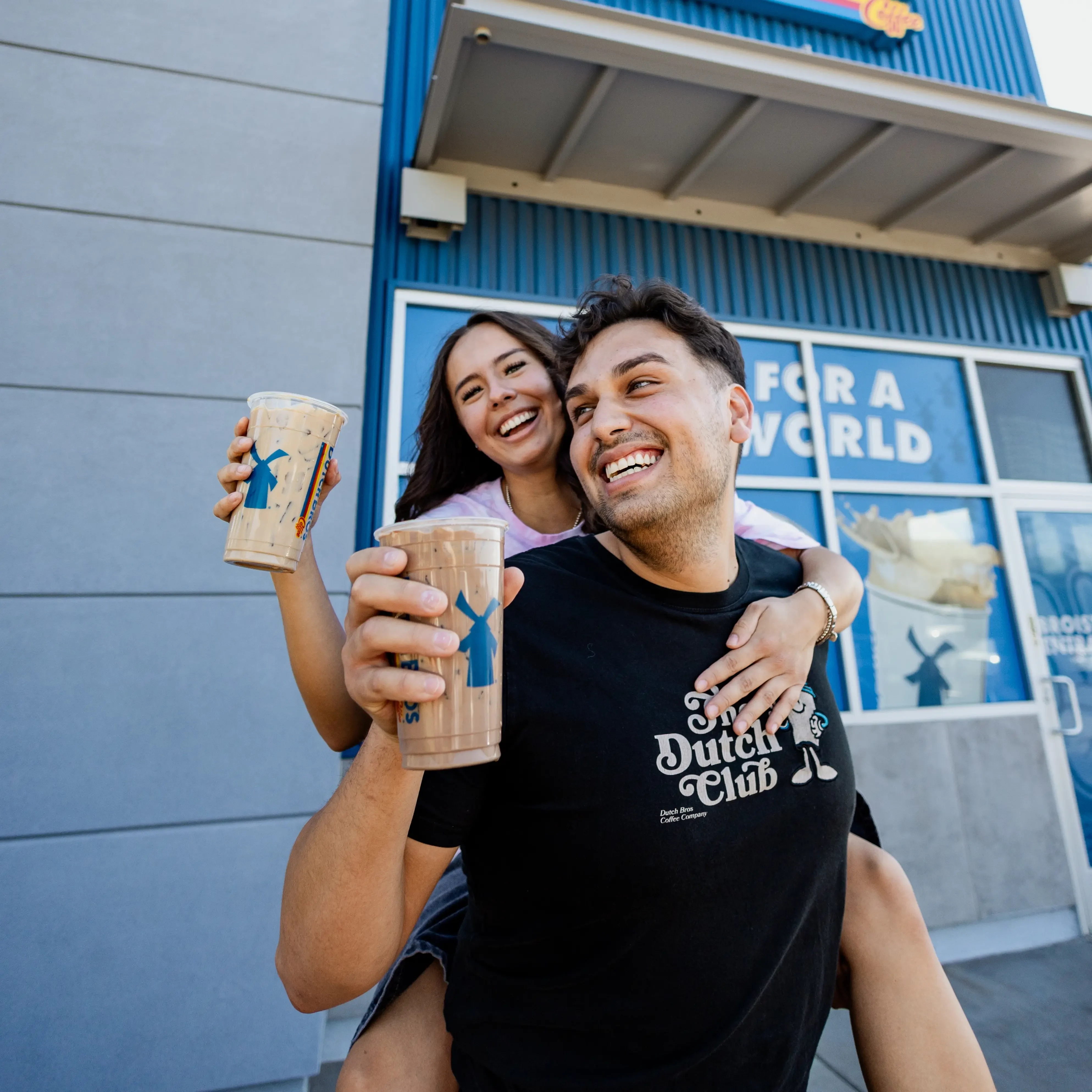 Two smiling people holding Dutch Bros coffee cups outside a blue storefront, one wearing a Dutch Club t-shirt.