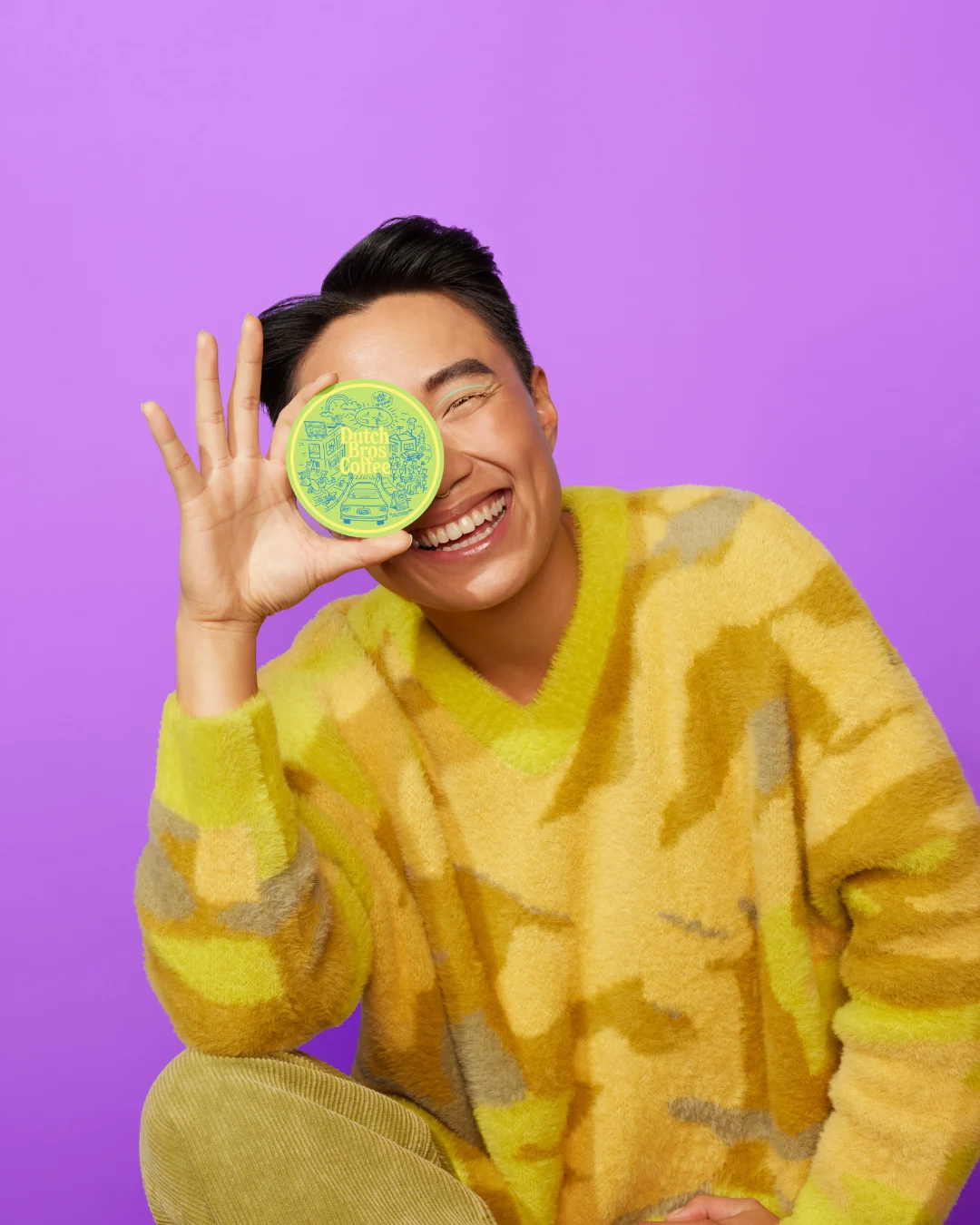 Person in yellow sweater holding a green "Dutch Bros Coffee" container over one eye against purple background.
