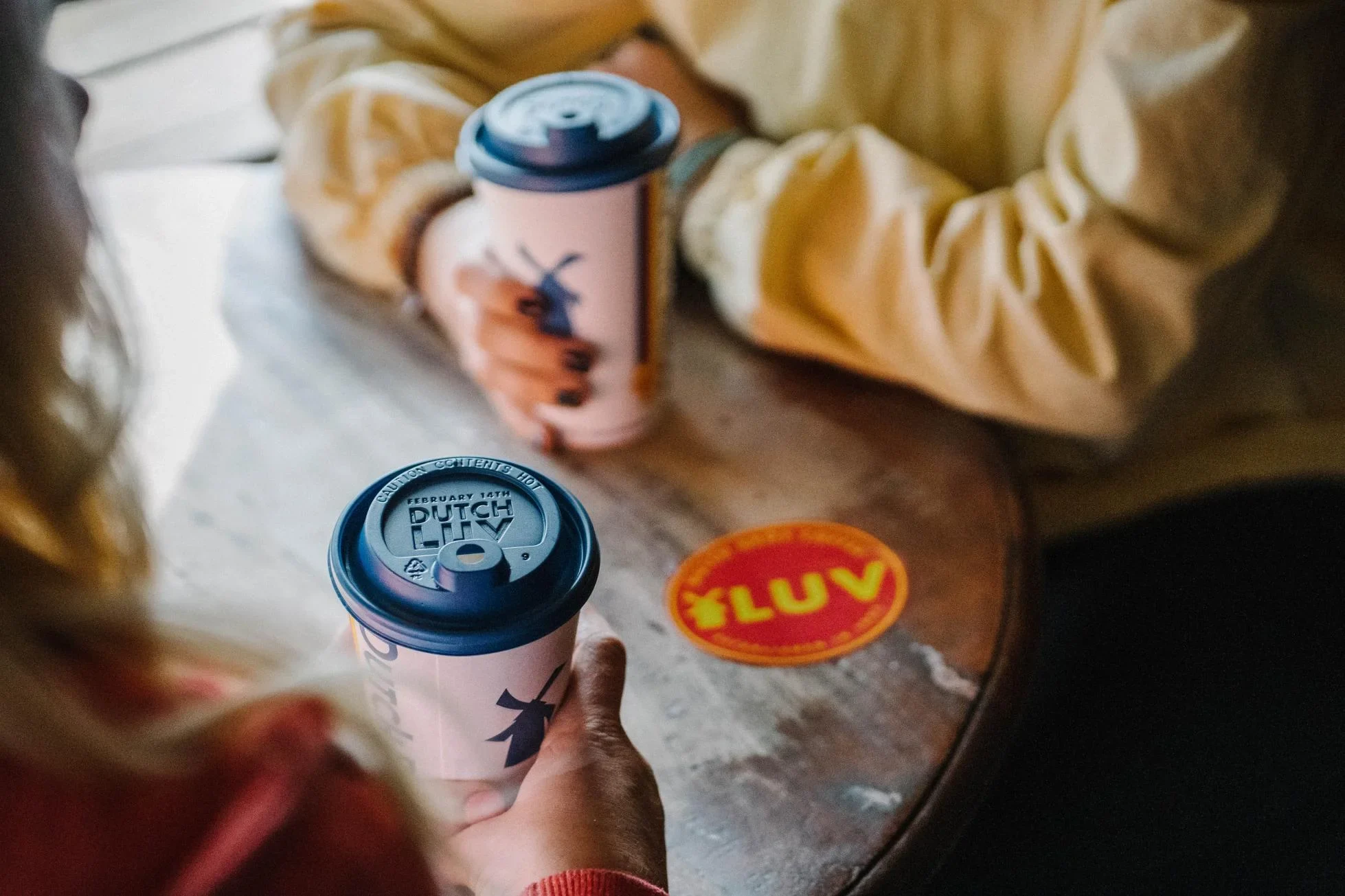 Two Dutch Bros coffee cups with blue lids held by hands, with a yellow and red "ILUV" sticker visible on the table.