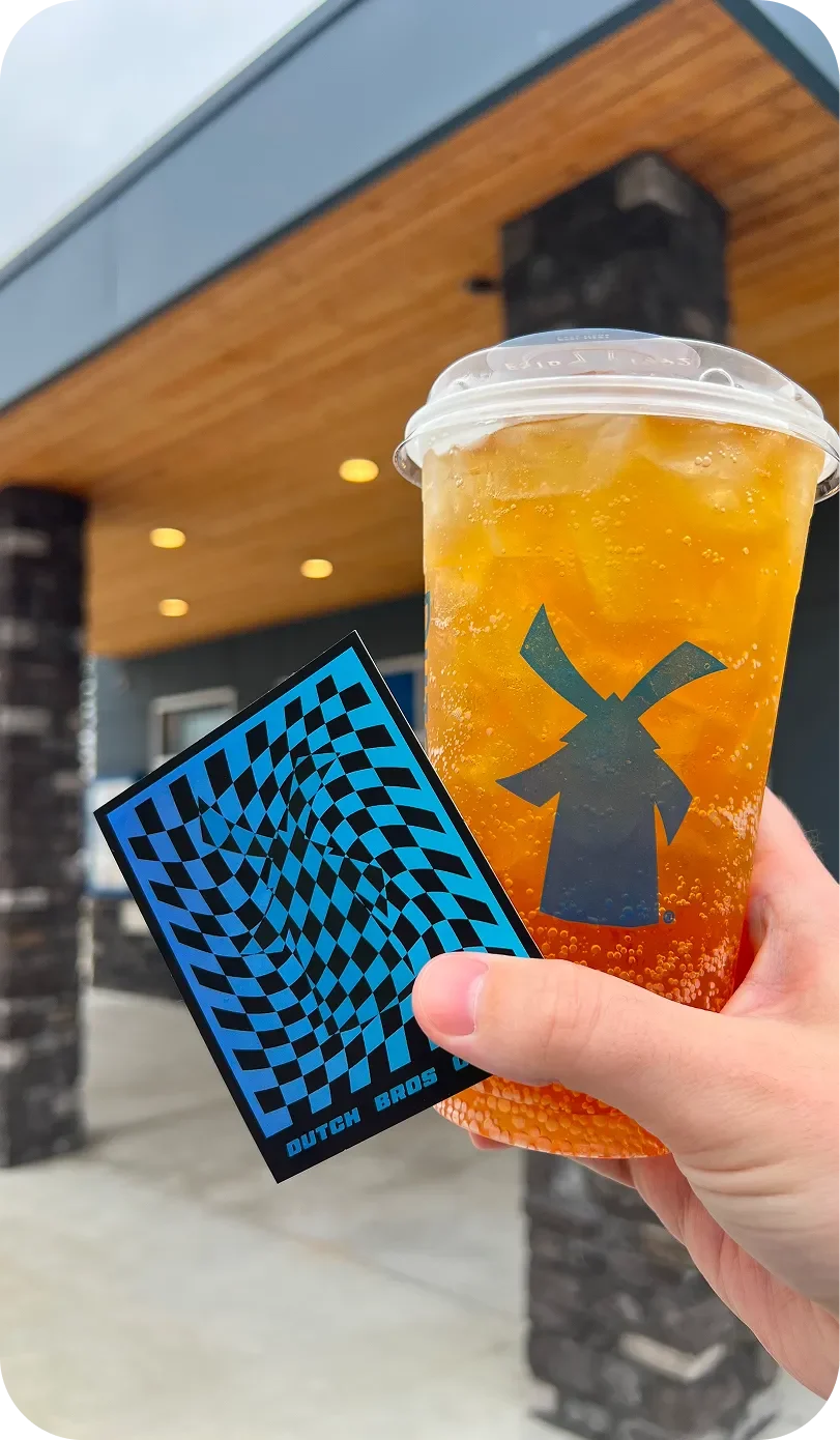 A hand holds a Dutch Bros iced drink and a blue checkered sticker outside a Dutch Bros coffee shop.