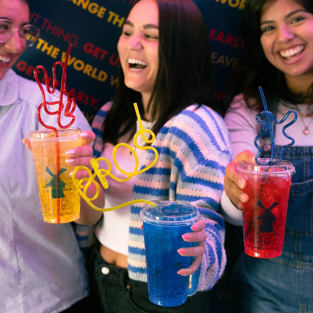 Three smiling friends holding colorful drinks with fun shaped straws in yellow, red and blue against a dark background.
