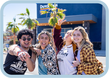 Four young friends posing together outdoors on a sunny day with palm trees and a storefront in the background.