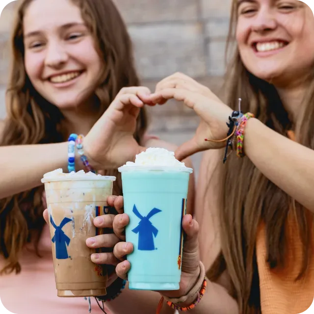 Two smiling girls holding Dutch Bros drinks topped with whipped cream, making a heart shape with their hands.