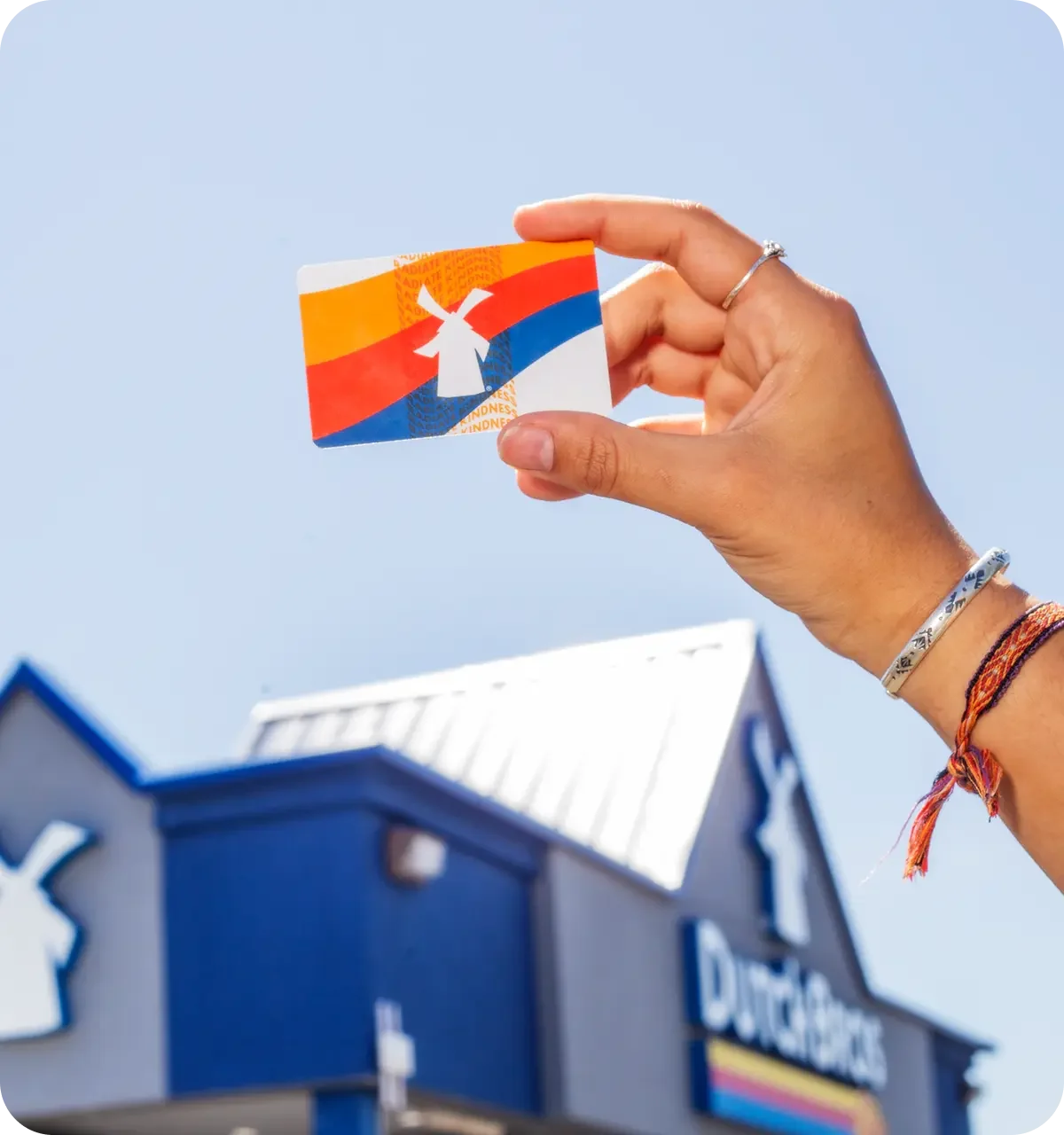 Hand holding a colorful Dutch gift card with windmill design against blue sky, with store building in background.