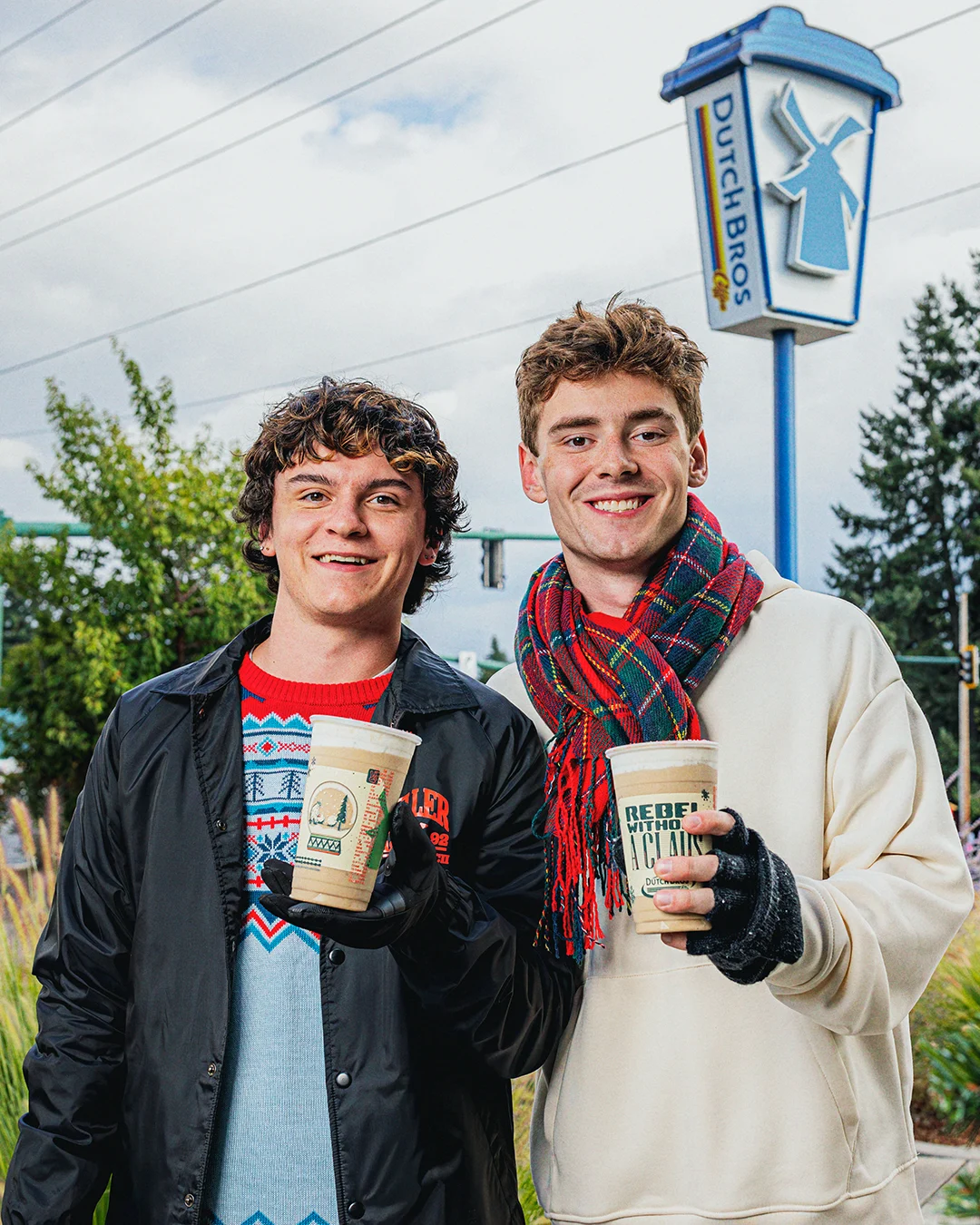 Two young men holding coffee cups outside a Dutch Bros coffee shop with the windmill sign visible above.