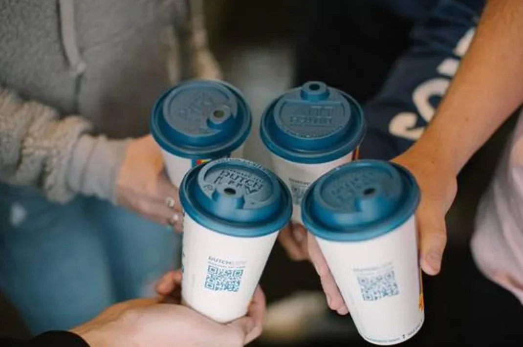 Hands holding four takeaway coffee cups with blue lids and QR codes, toasting together in a social gathering.