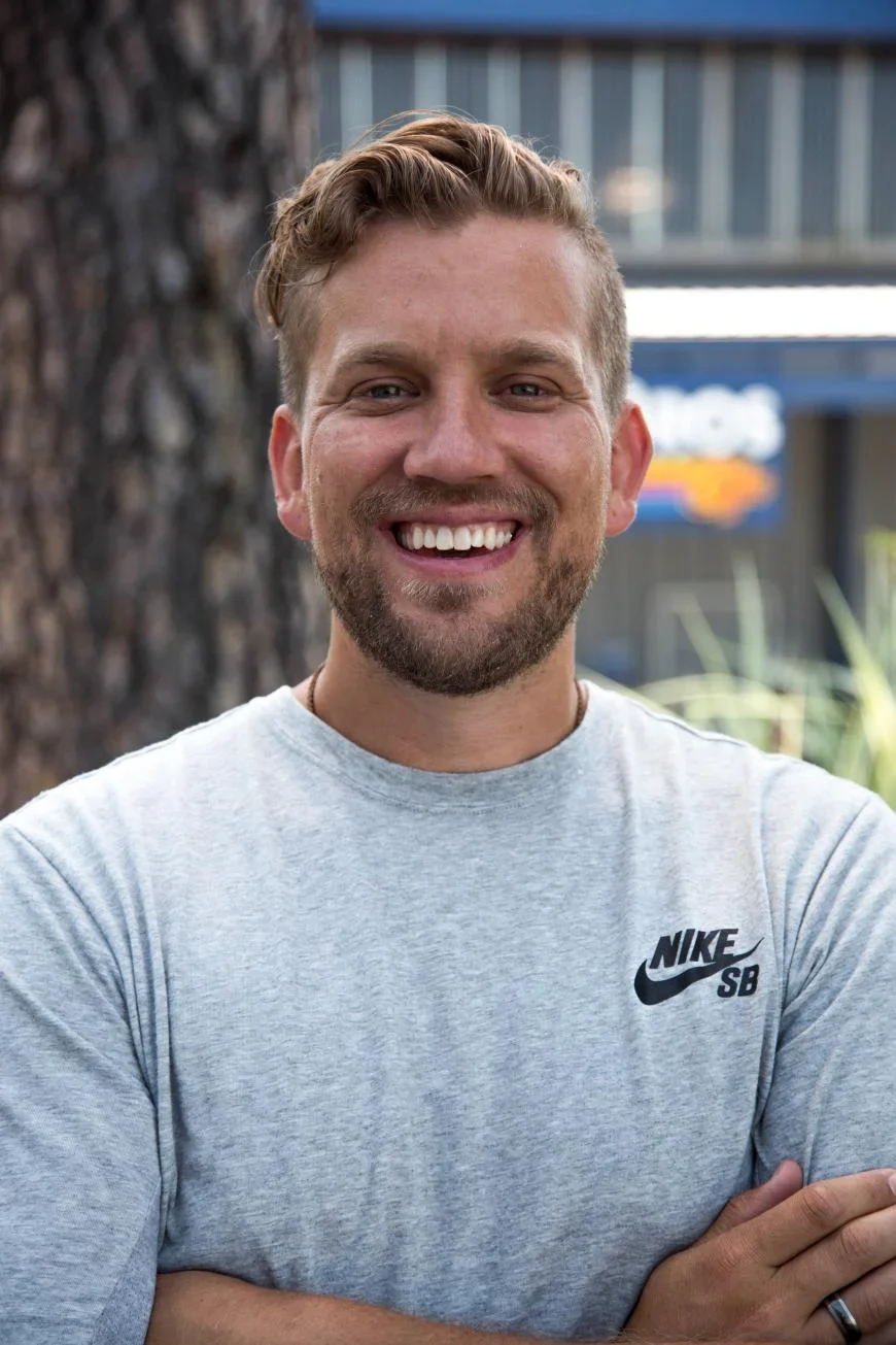 Person smiling outdoors wearing a light gray Nike SB t-shirt with arms crossed, blurred building in background.