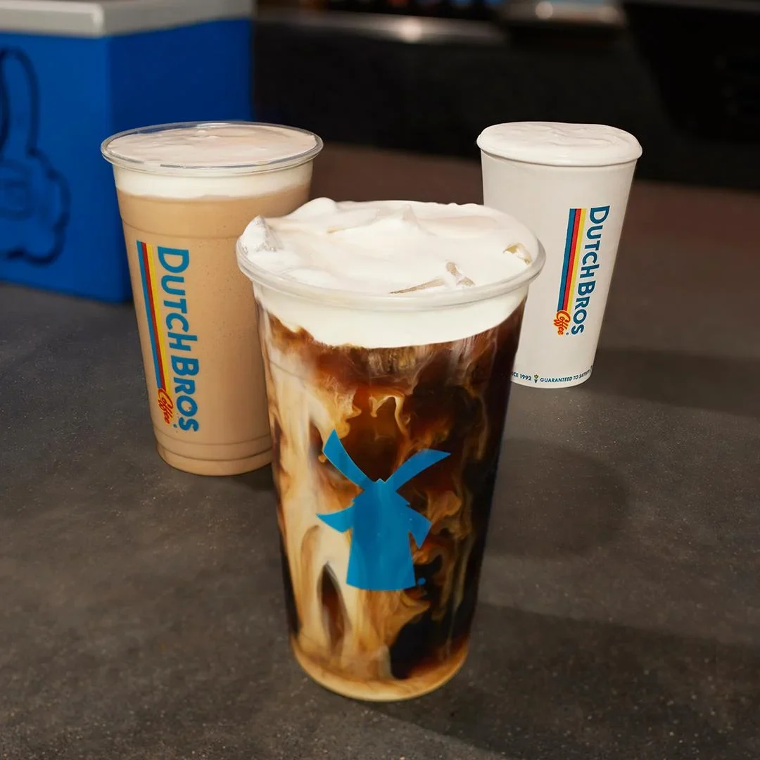 Three Dutch Bros coffee drinks on a dark counter - a creamy iced coffee, a marbled drink with whipped cream, and a white cup.