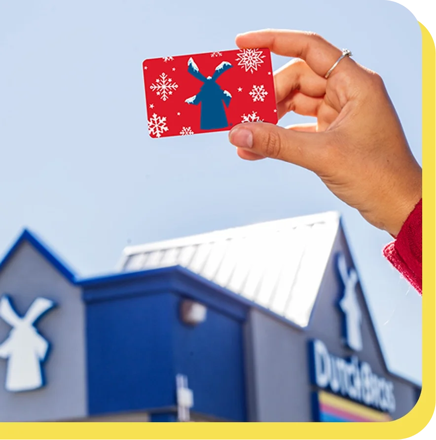Hand holding a red gift card with blue windmill and snowflakes in front of a Dutch Bros coffee shop building.