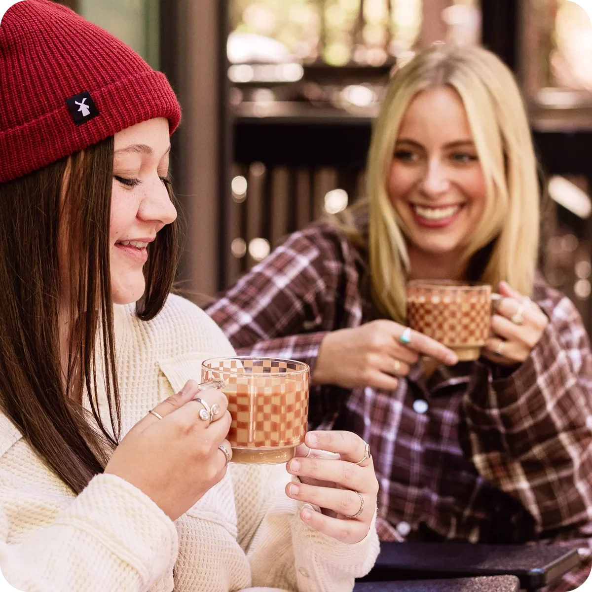 Two women smile and hold checkered glass mugs of coffee, one wearing a red beanie and white sweater, the other in a plaid shirt.