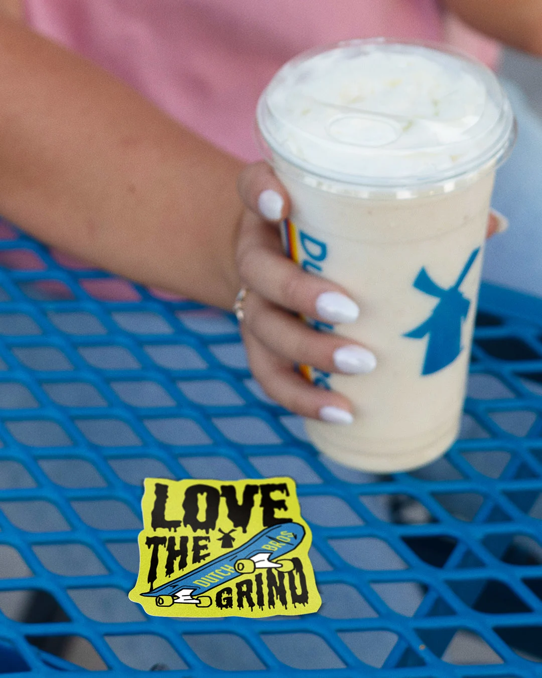 Hand with white nails holding an iced coffee drink on blue metal table with "LOVE THE GRIND" skateboard sticker.
