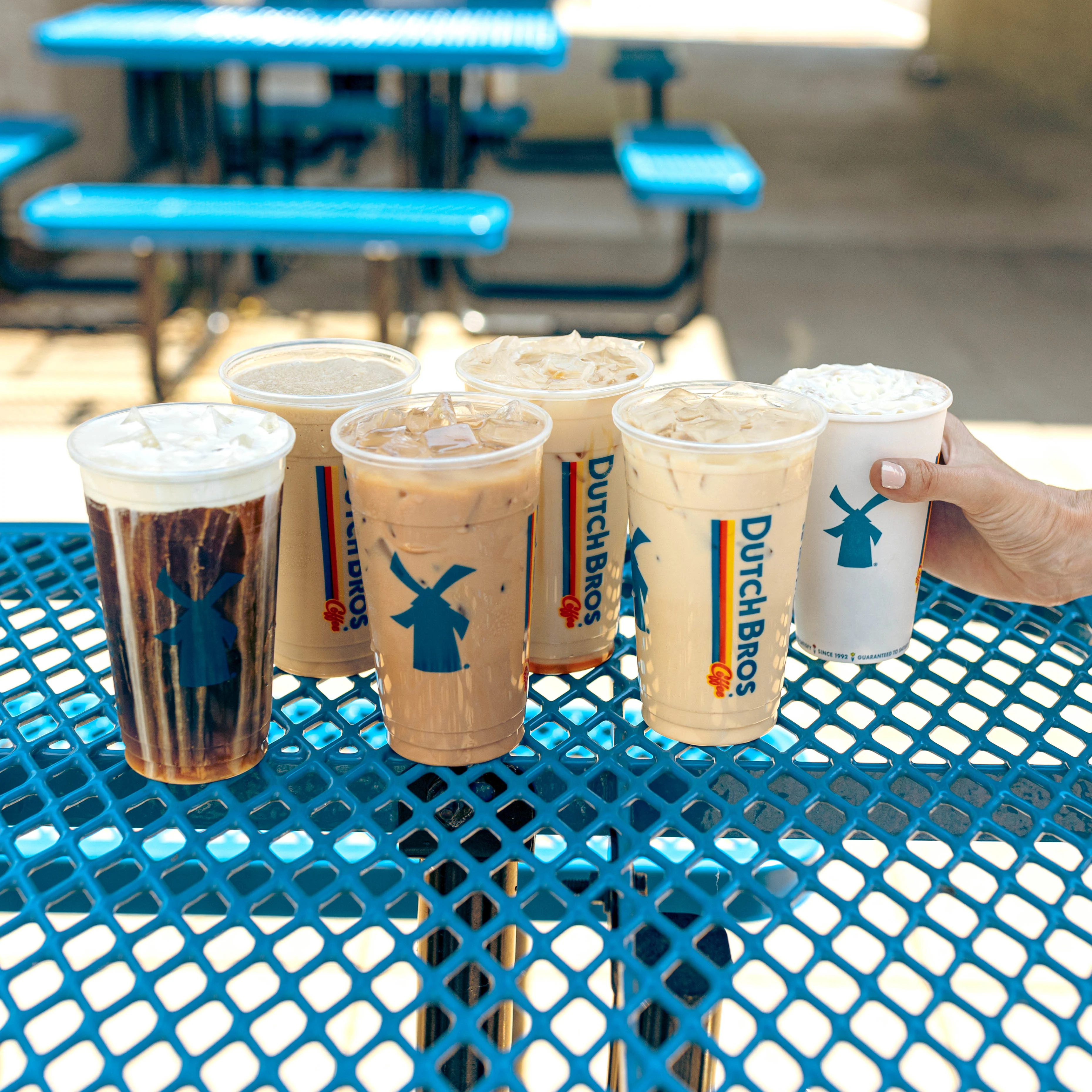 Assorted Dutch Bros coffee drinks on blue metal table, with hand reaching for rightmost cup, outdoor seating visible behind.
