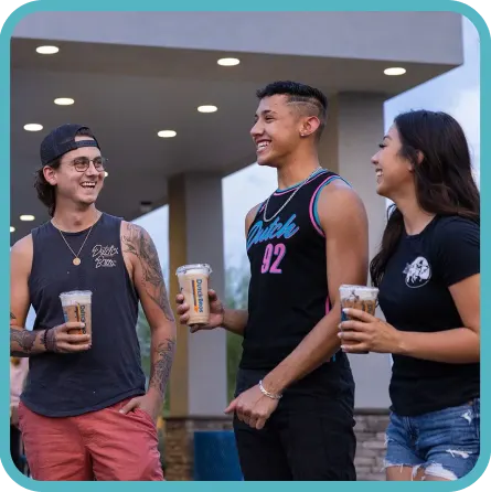 Three young people smiling and talking while holding drinks outside a building with recessed lighting.