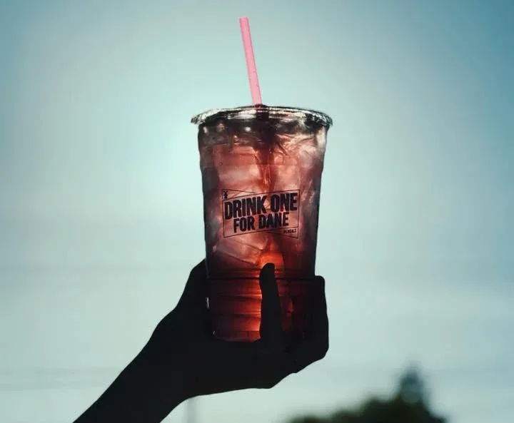 Hand holding a clear plastic cup with red drink and ice, labeled "DRINK ONE FOR DANE" with a pink straw against blue sky.