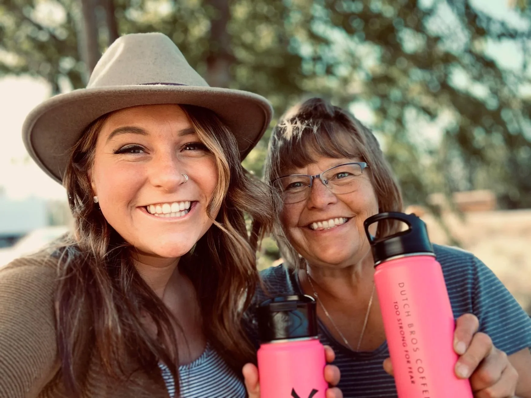 Two smiling women outdoors, one wearing a beige hat, both holding pink water bottles against a blurred green background.