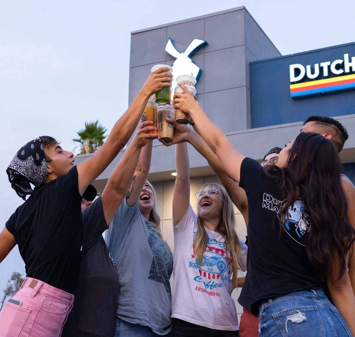 Group of friends toasting with Dutch Bros coffee drinks outside store with windmill logo visible in background.