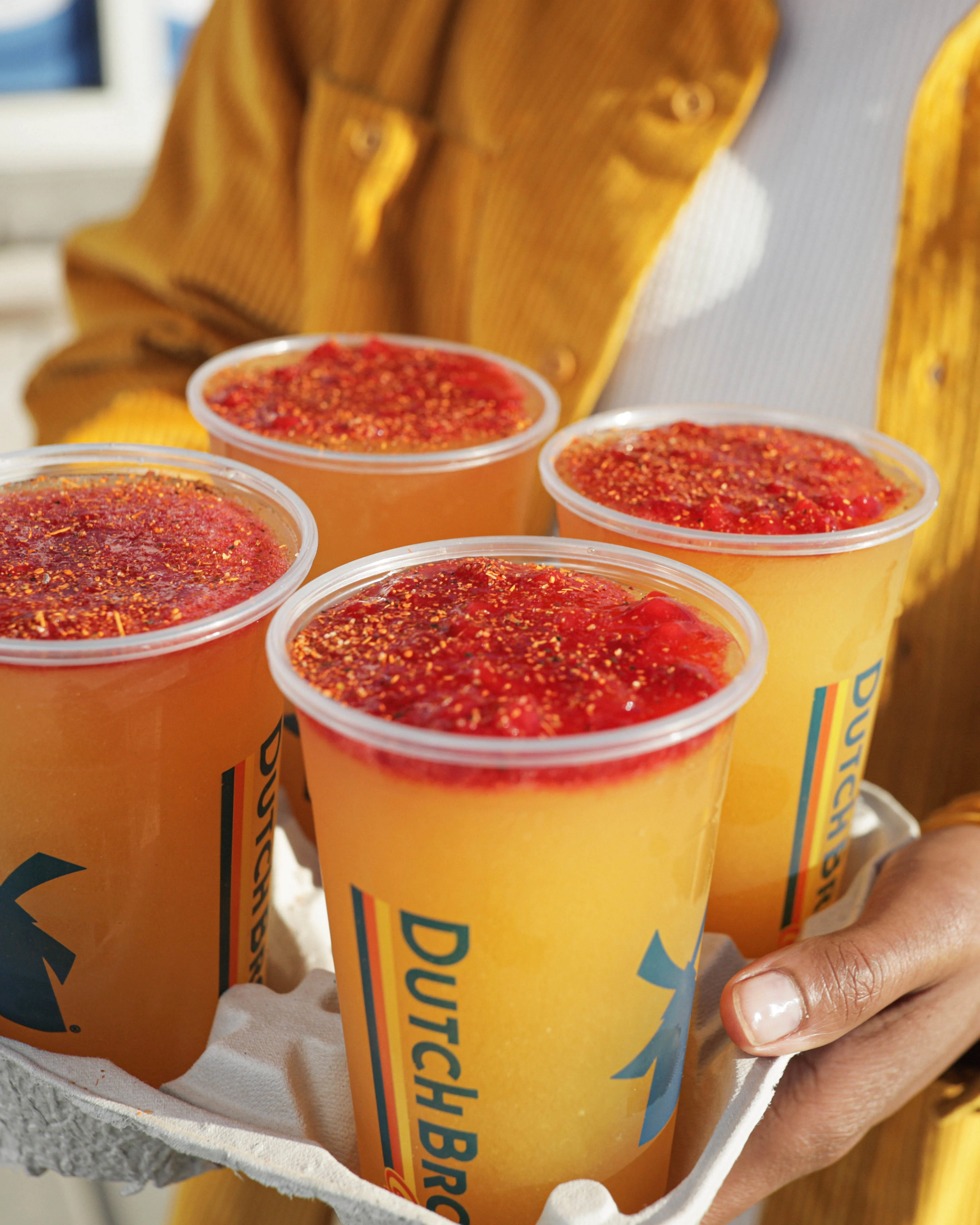 Four Dutch Bros drinks in a tray with yellow mango base and red topping, held by someone in a mustard sweater.