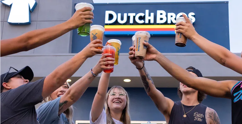 Group toasting with colorful drinks outside Dutch Bros coffee shop, raising cups in celebration.
