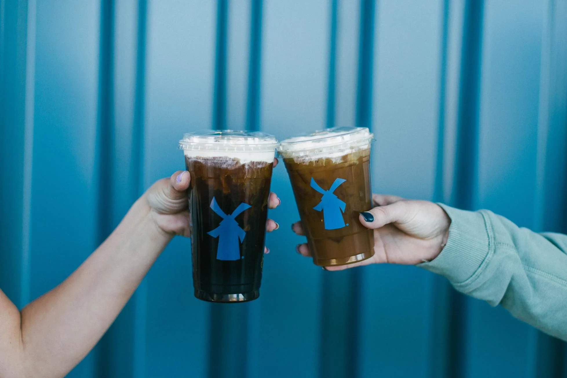 Two hands toasting iced coffee drinks with blue windmill logos against a teal blue background.