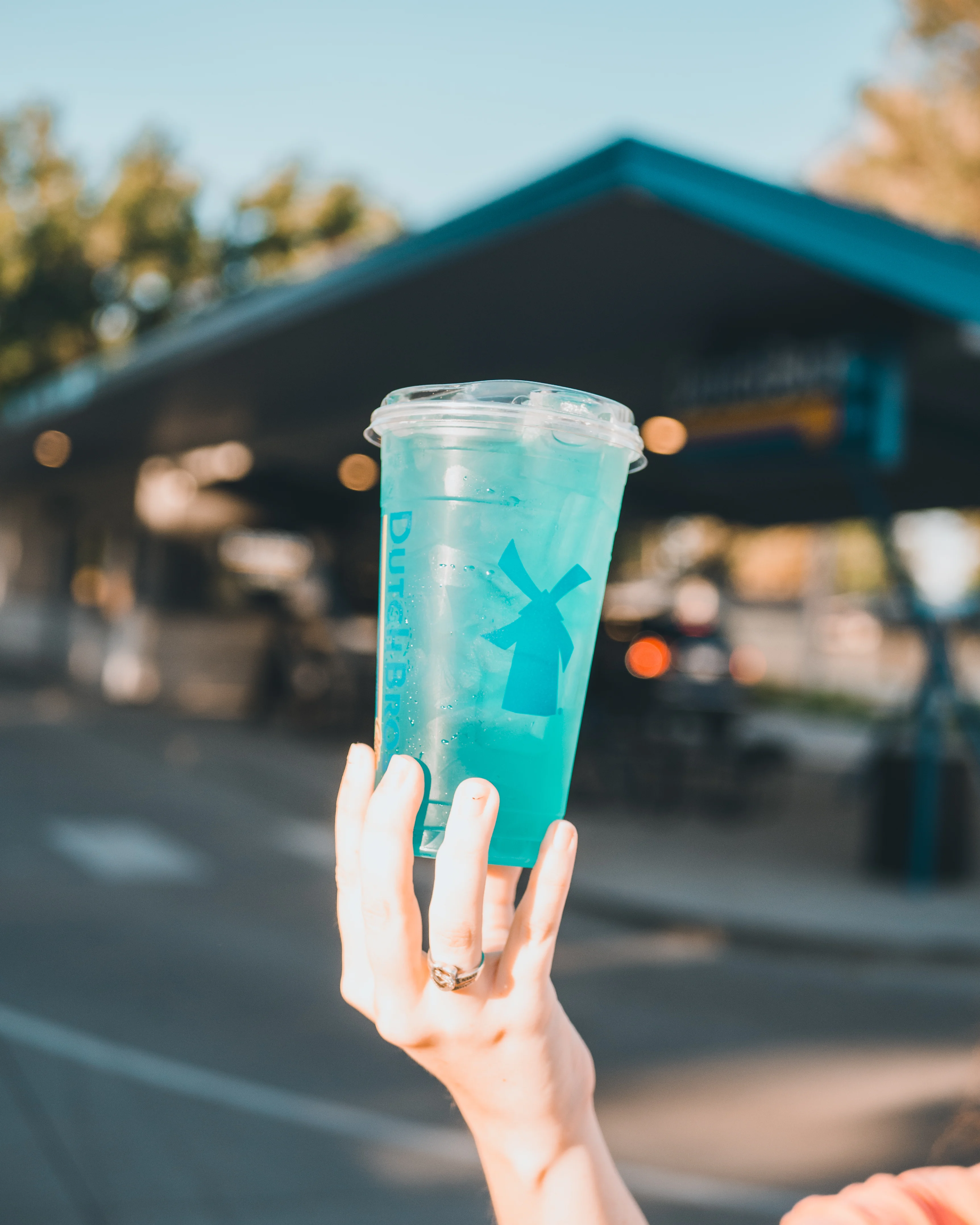 Hand holding a bright blue Dutch Bros iced drink in a clear cup with windmill logo against a cafe backdrop.