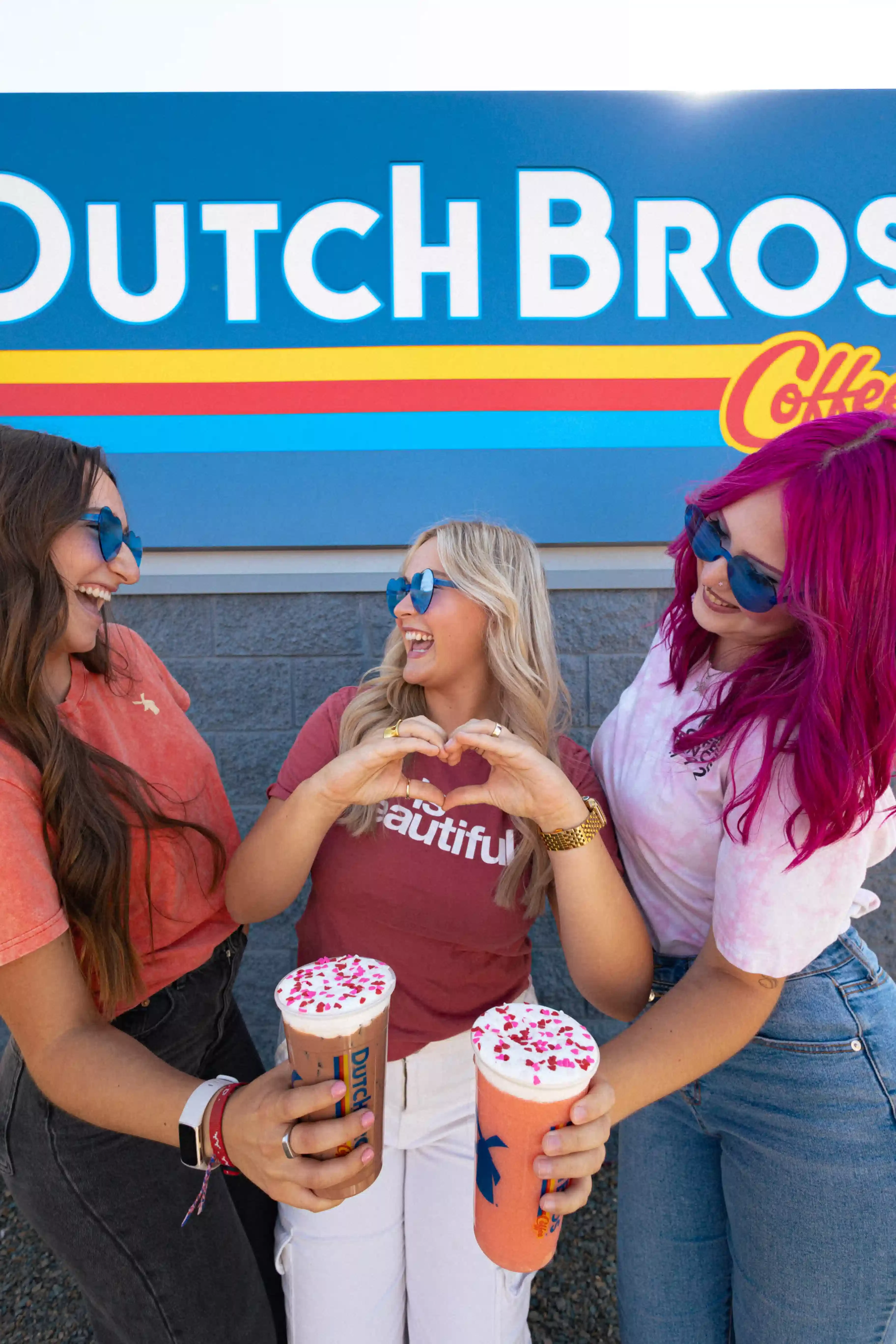 Three friends in blue sunglasses enjoying colorful drinks outside Dutch Bros Coffee, one making heart gesture.