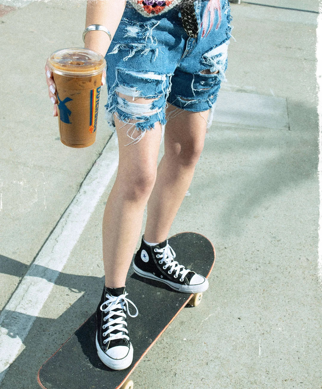 Person on skateboard wearing distressed denim shorts and black Converse sneakers, holding iced coffee.
